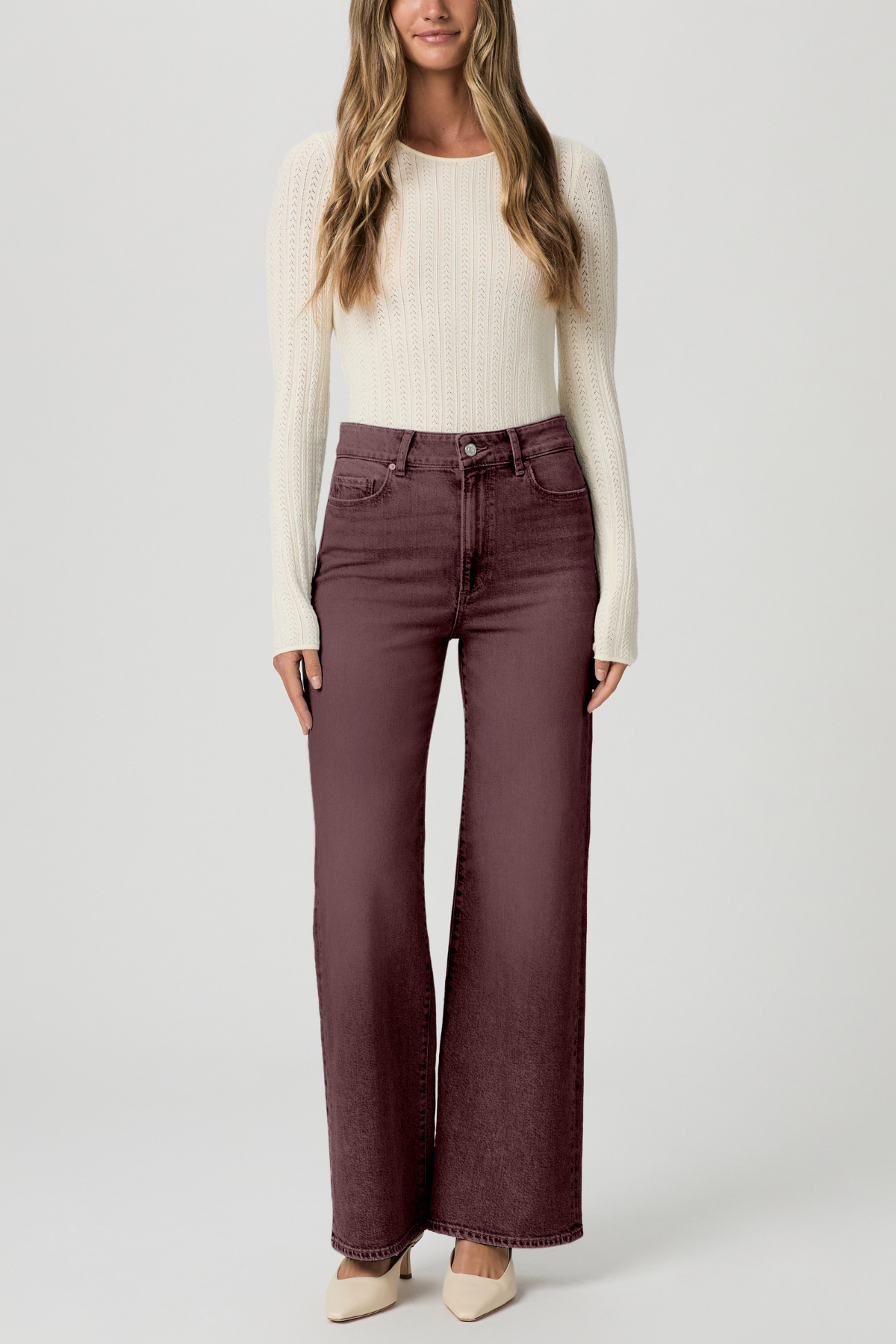 A woman modeling a pair of high-waisted, wide-leg jeans in a dusty plum purple color, paired with a cream ribbed knit sweater and beige pointed-toe heels, against a light gray studio background.