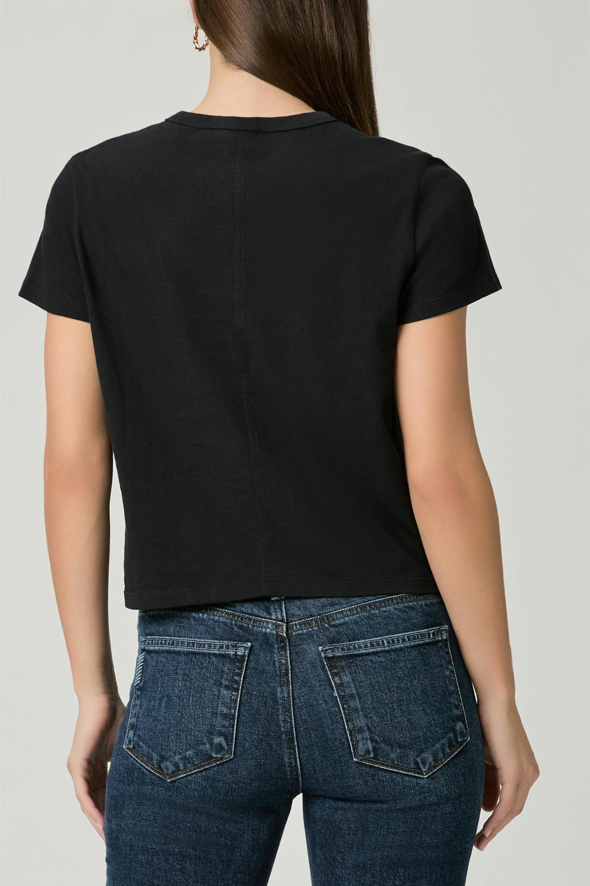 A woman viewed from the back wearing a black short-sleeve crop top and dark blue jeans against a light gray studio background.
