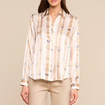 A woman modeling a long-sleeve button-up blouse with vertical beige and white stripes and small blue circular accents, paired with beige pants, against a solid beige studio background.