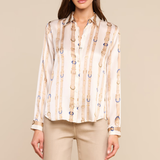 A woman modeling a long-sleeve button-up blouse with vertical beige and white stripes and small blue circular accents, paired with beige pants, against a solid beige studio background.