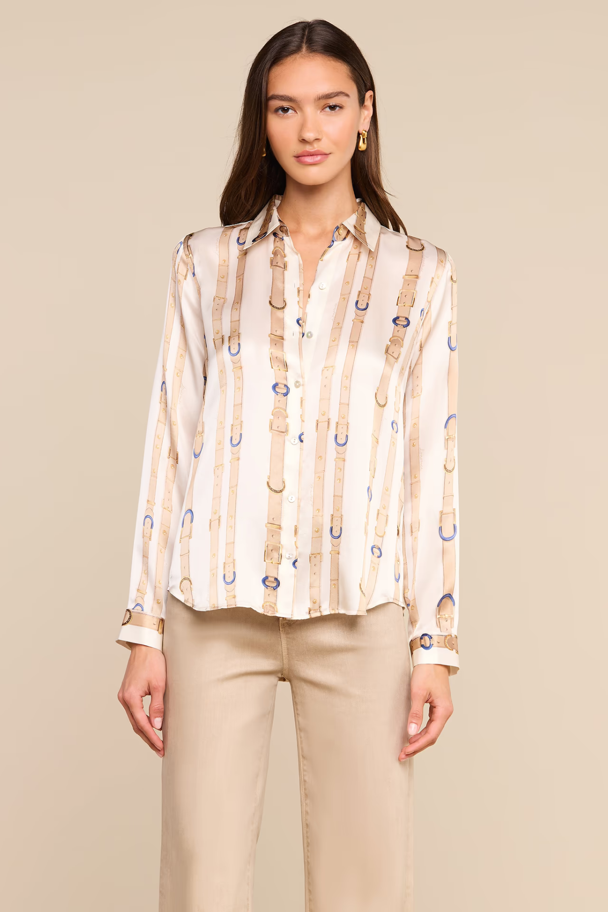 A woman modeling a long-sleeve button-up blouse with vertical beige and white stripes and small blue circular accents, paired with beige pants, against a solid beige studio background.