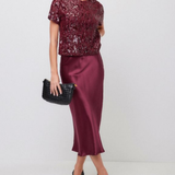 A woman models a two-piece outfit consisting of a short-sleeve burgundy sequined top and a matching satin midi skirt, holding a black clutch bag.