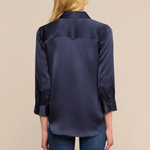 A woman viewed from the back wearing a dark navy satin button-up shirt and blue jeans, set against a solid beige studio background.