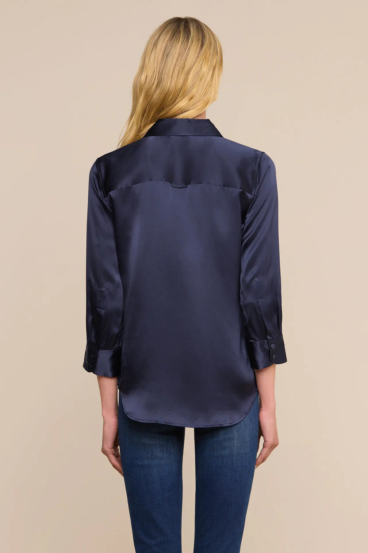 A woman viewed from the back wearing a dark navy satin button-up shirt and blue jeans, set against a solid beige studio background.