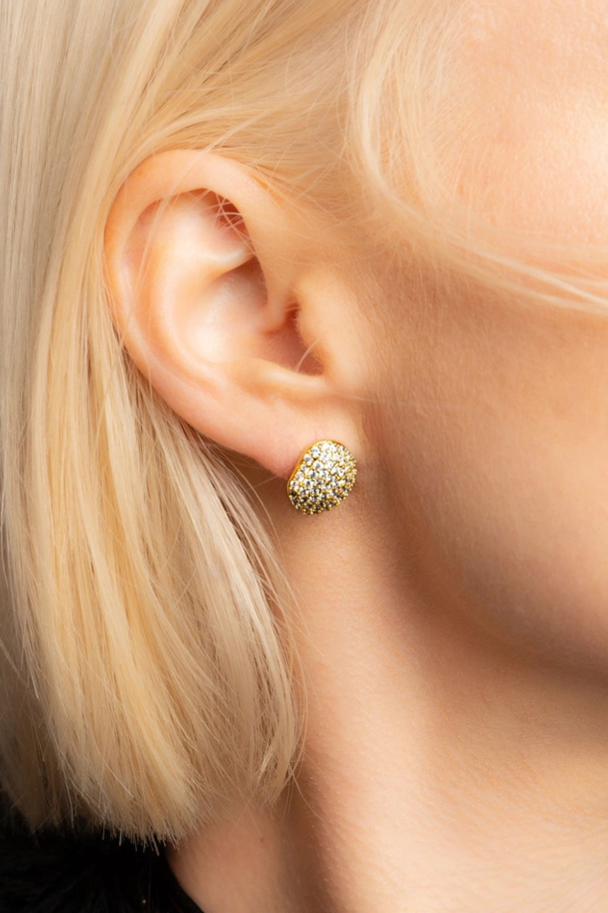 A close-up of a model's ear wearing a small gold stud earring encrusted with clear stones.