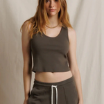 A woman modeling a dark brown cropped tank top and matching dark brown sweatpants with a white drawstring, set against a neutral draped fabric backdrop.