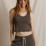 A woman modeling a dark brown cropped tank top and matching dark brown sweatpants with a white drawstring, set against a neutral draped fabric backdrop.