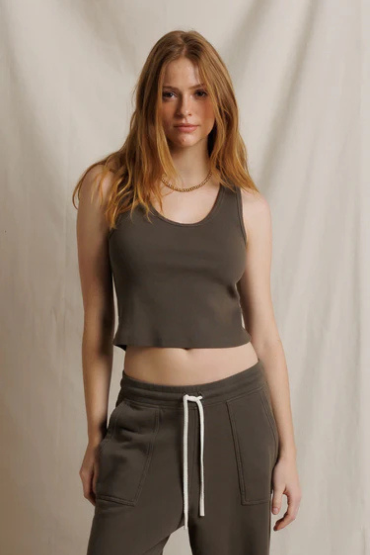 A woman modeling a dark brown cropped tank top and matching dark brown sweatpants with a white drawstring, set against a neutral draped fabric backdrop.