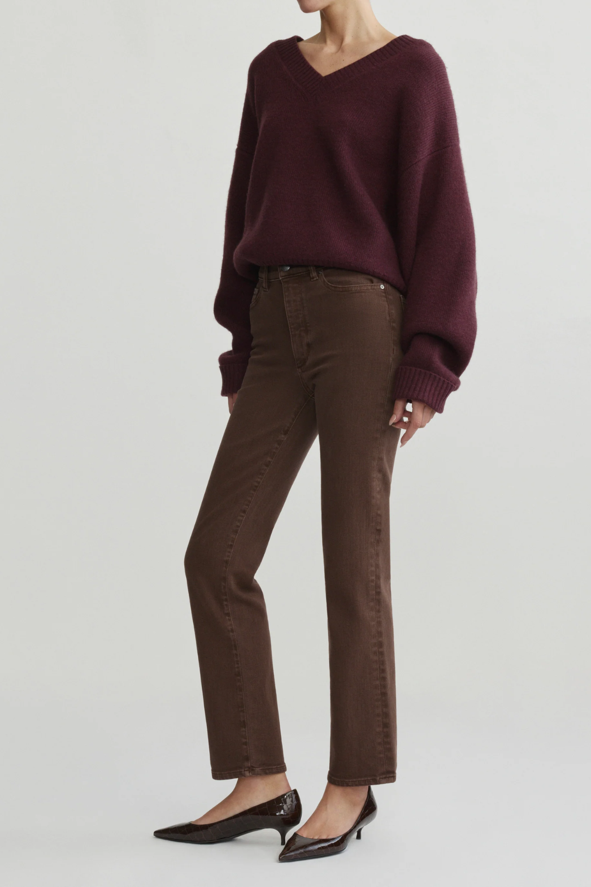 A model wearing a dark burgundy knit V-neck sweater, brown straight-leg jeans, and black pointed-toe flats, photographed against a light gray studio background.