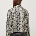 A woman viewed from the back wearing a cropped long-sleeve jacket with a snakeskin pattern in beige, gray, and black, paired with black leather pants, against a light gray studio background.