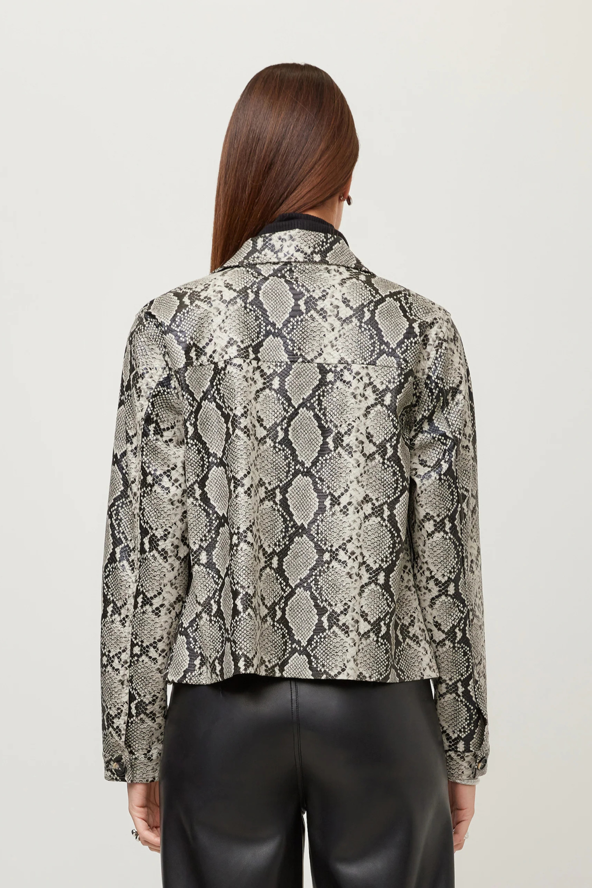 A woman viewed from the back wearing a cropped long-sleeve jacket with a snakeskin pattern in beige, gray, and black, paired with black leather pants, against a light gray studio background.