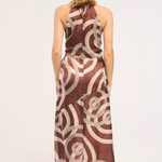 A woman viewed from behind wearing a sleeveless halter-neck maxi dress with a brown and beige abstract circular pattern, standing against a plain light background.