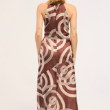 A woman viewed from behind wearing a sleeveless halter-neck maxi dress with a brown and beige abstract circular pattern, standing against a plain light background.