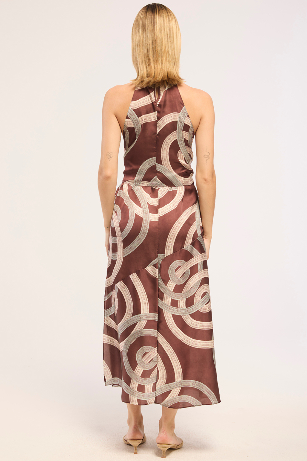A woman viewed from behind wearing a sleeveless halter-neck maxi dress with a brown and beige abstract circular pattern, standing against a plain light background.