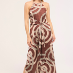 A woman modeling a sleeveless halter-neck midi dress with a brown and beige geometric spiral pattern, standing against a plain light background.