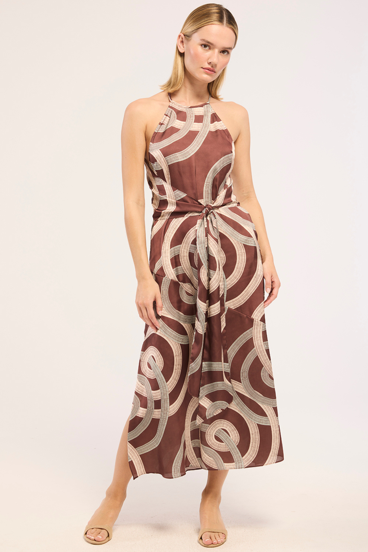 A woman modeling a sleeveless halter-neck midi dress with a brown and beige geometric spiral pattern, standing against a plain light background.