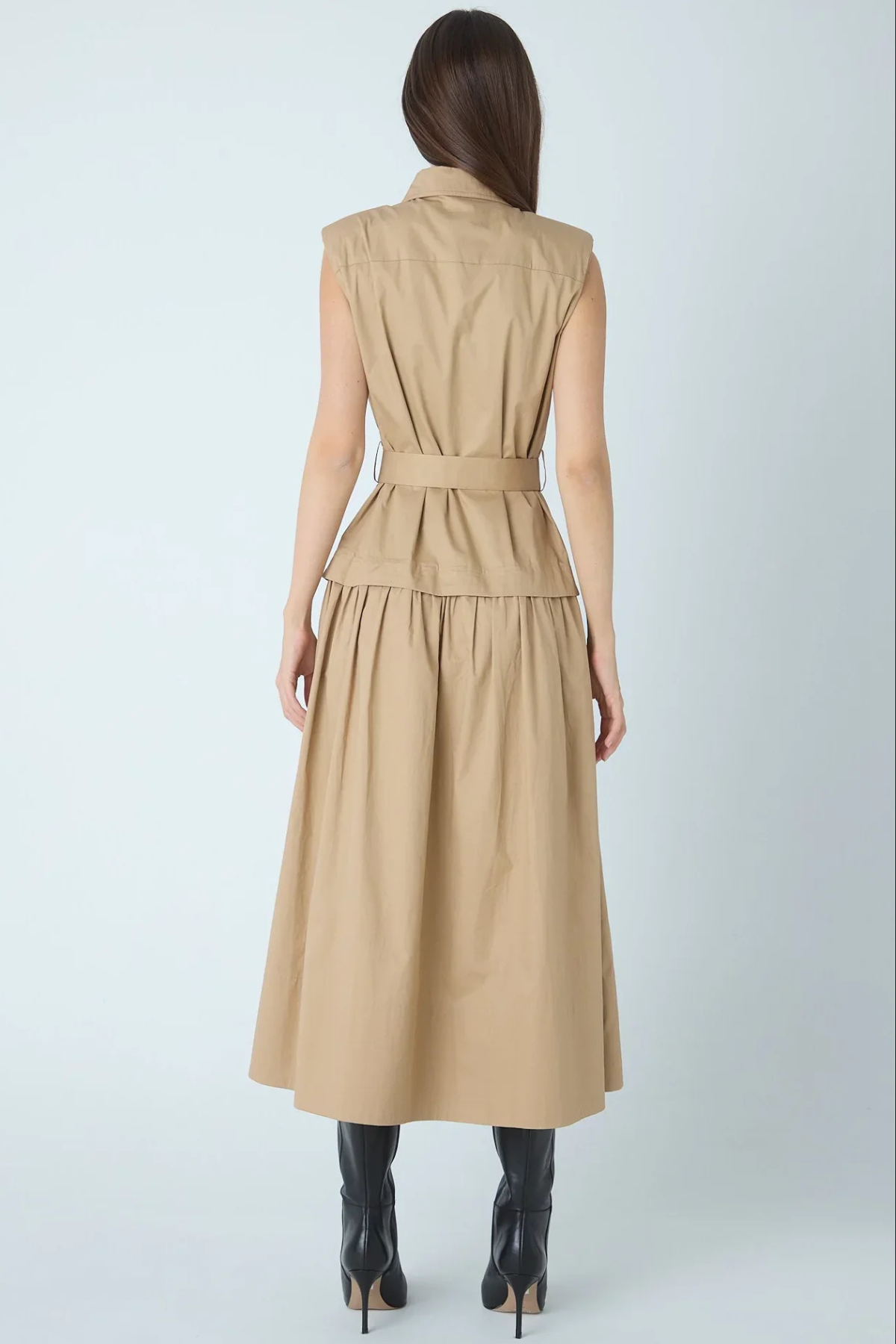A woman shown from the back wearing a sleeveless beige midi dress with a defined waist and a flared skirt, paired with black ankle boots, against a light gray studio background.