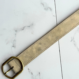 A light beige suede belt with a prominent gold-tone metal buckle on a marble background