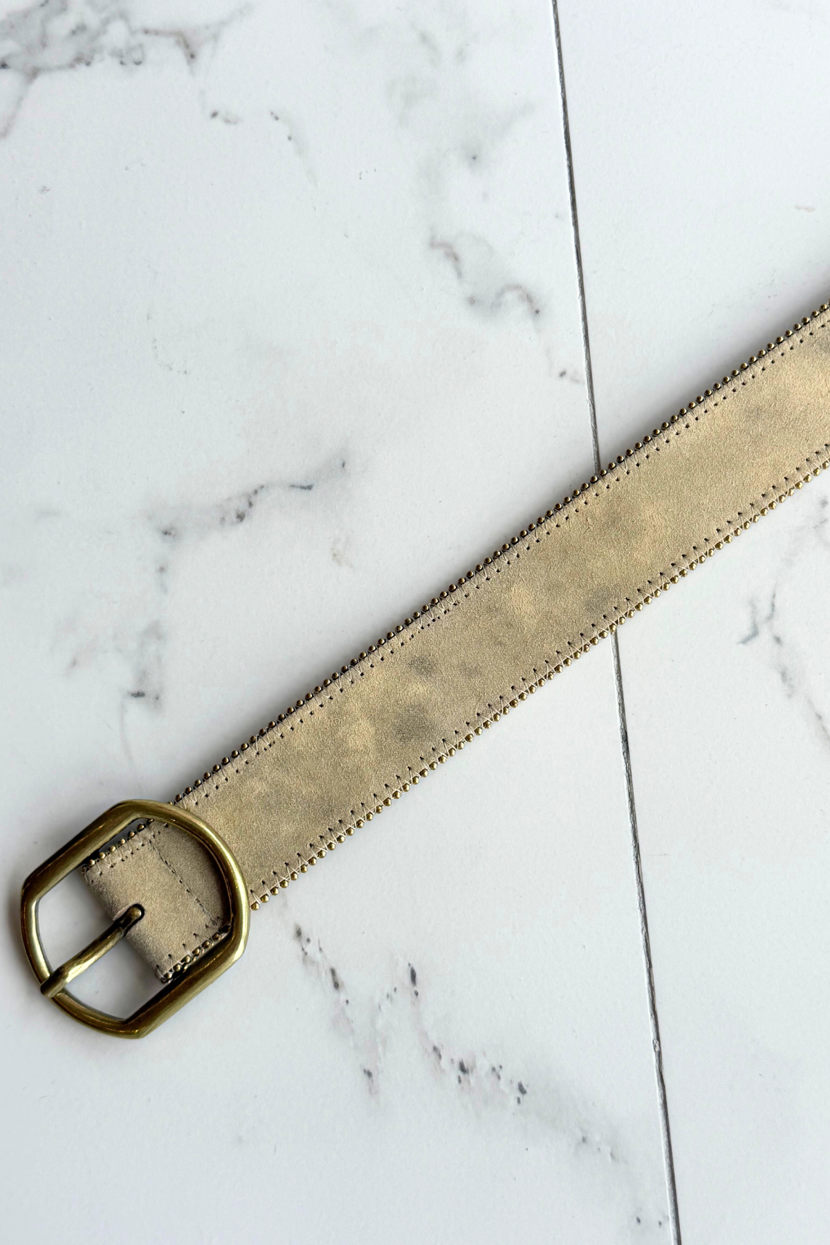 A light beige suede belt with a prominent gold-tone metal buckle on a marble background
