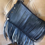A black leather fringe clutch bag with a top zipper and a single shoulder strap, resting on a beige tufted leather chair.