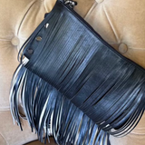 A black leather fringe clutch bag with a top zipper and a single shoulder strap, resting on a beige tufted leather chair.
