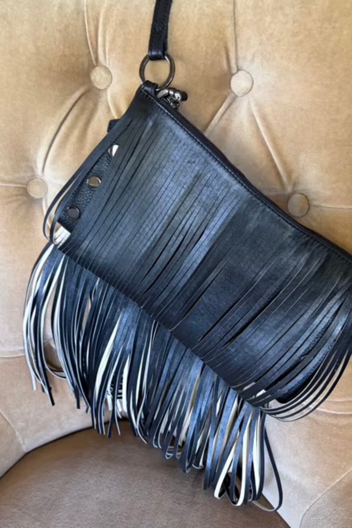 A black leather fringe clutch bag with a top zipper and a single shoulder strap, resting on a beige tufted leather chair.
