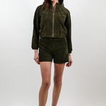 A woman modeling a dark olive green two-piece set consisting of a cropped zip-up jacket and matching shorts, paired with white pointed-toe heels, against a light gray studio background.