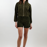 A woman modeling a dark olive green two-piece set consisting of a cropped zip-up jacket and matching shorts, paired with white pointed-toe heels, against a light gray studio background.