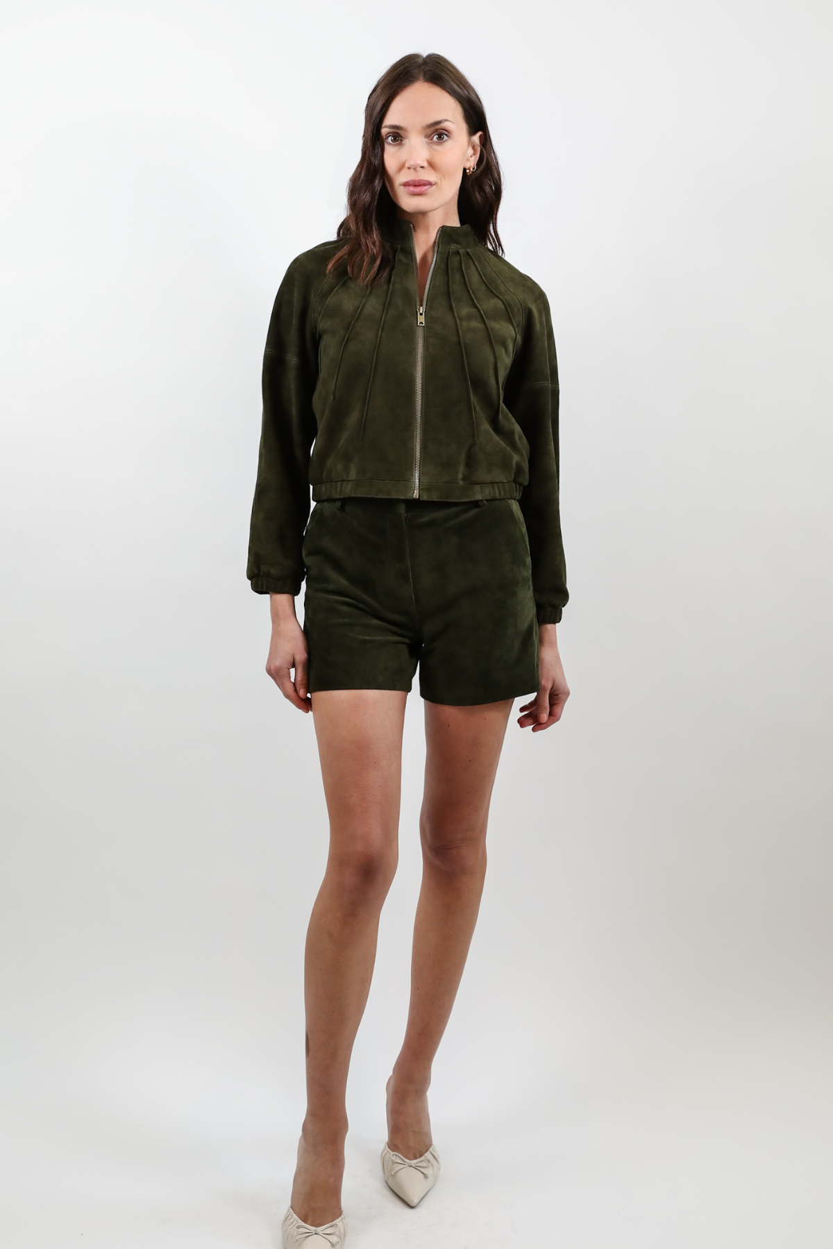 A woman modeling a dark olive green two-piece set consisting of a cropped zip-up jacket and matching shorts, paired with white pointed-toe heels, against a light gray studio background.