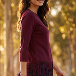A woman wearing a dark burgundy long-sleeve top with fringe trim at the hem, paired with dark blue jeans, standing outdoors in a sunlit forest setting.