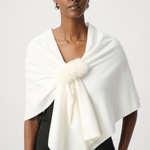 A model wearing a cream-colored wrap shawl with a fluffy pom-pom detail at the center, styled over a black top against a light gray studio background.