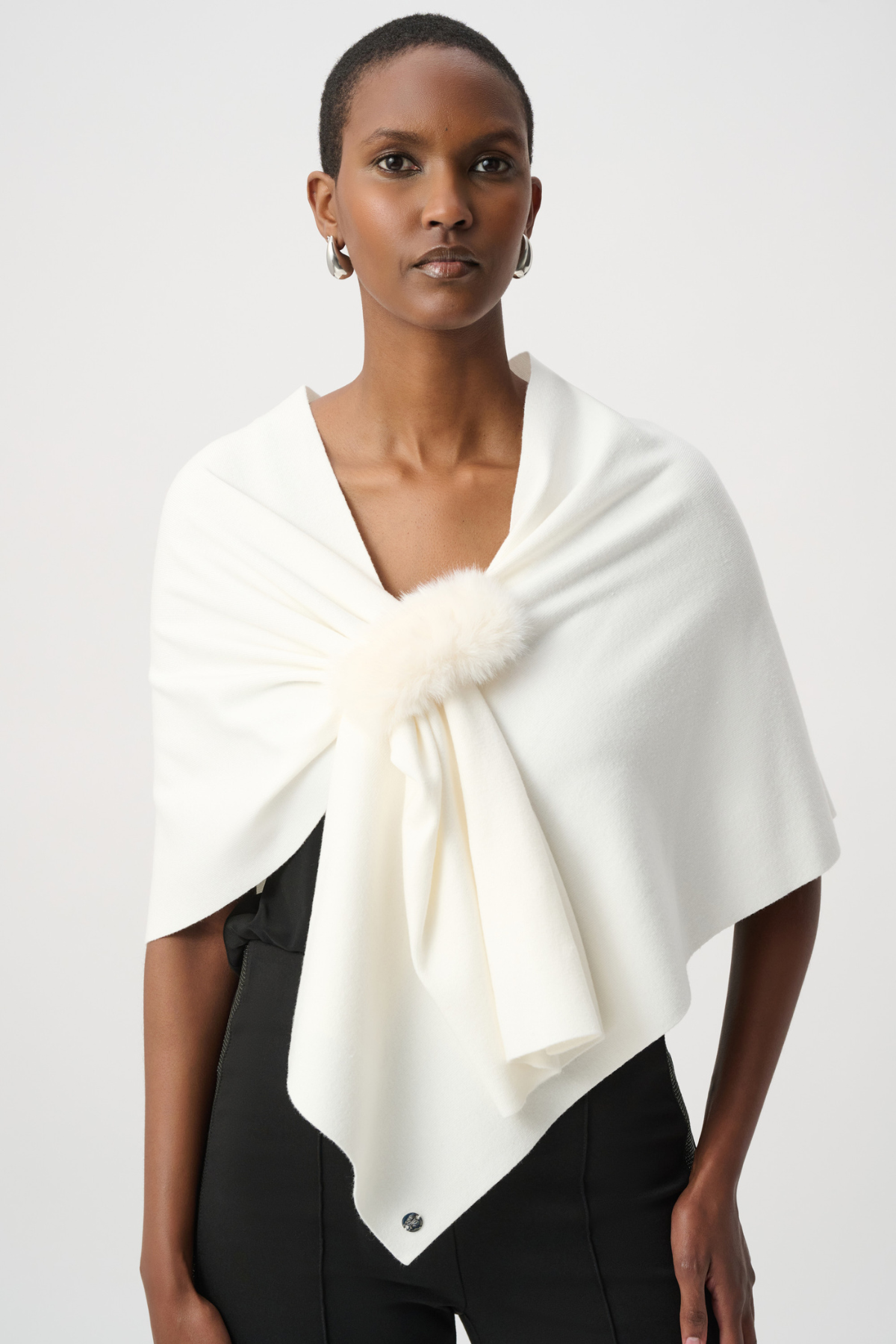 A model wearing a cream-colored wrap shawl with a fluffy pom-pom detail at the center, styled over a black top against a light gray studio background.