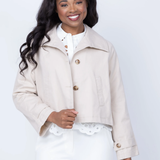 A smiling woman with long dark wavy hair models a light beige cropped jacket over a white top and white pants against a plain light gray studio background.