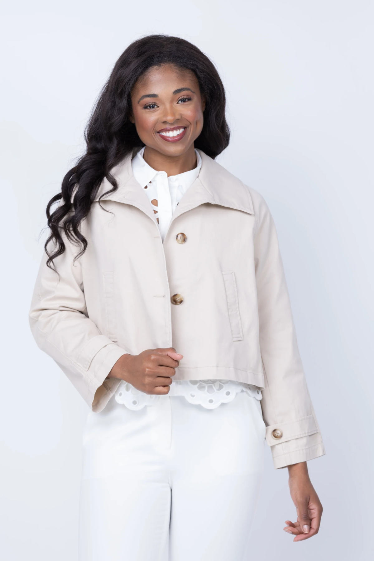 A smiling woman with long dark wavy hair models a light beige cropped jacket over a white top and white pants against a plain light gray studio background.