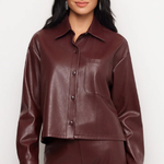 A woman modeling a cropped, long-sleeve faux leather button-up shirt in a deep burgundy color, paired with matching pants against a plain light gray background.