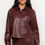 A woman modeling a cropped, long-sleeve faux leather button-up shirt in a deep burgundy color, paired with matching pants against a plain light gray background.
