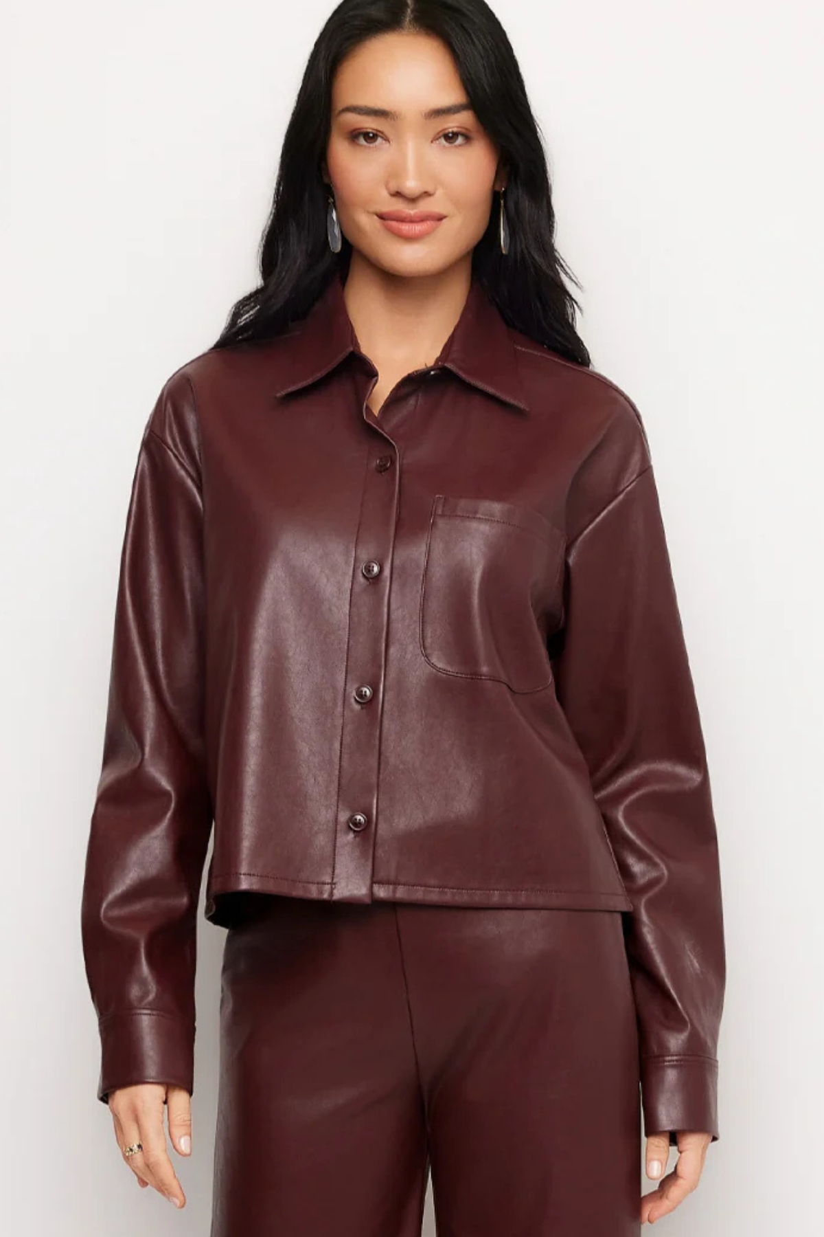 A woman modeling a cropped, long-sleeve faux leather button-up shirt in a deep burgundy color, paired with matching pants against a plain light gray background.
