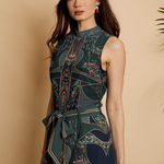 A woman modeling a sleeveless high-neck dress with a dark teal and navy floral print, featuring geometric patterns and a matching waist tie, set against a warm beige background.