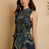 A woman modeling a sleeveless high-neck dress with a dark teal and navy floral print, featuring geometric patterns and a matching waist tie, set against a warm beige background.