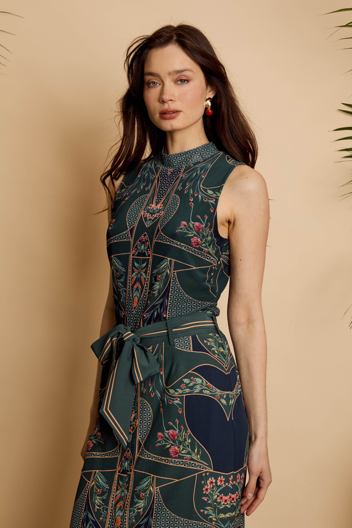A woman modeling a sleeveless high-neck dress with a dark teal and navy floral print, featuring geometric patterns and a matching waist tie, set against a warm beige background.