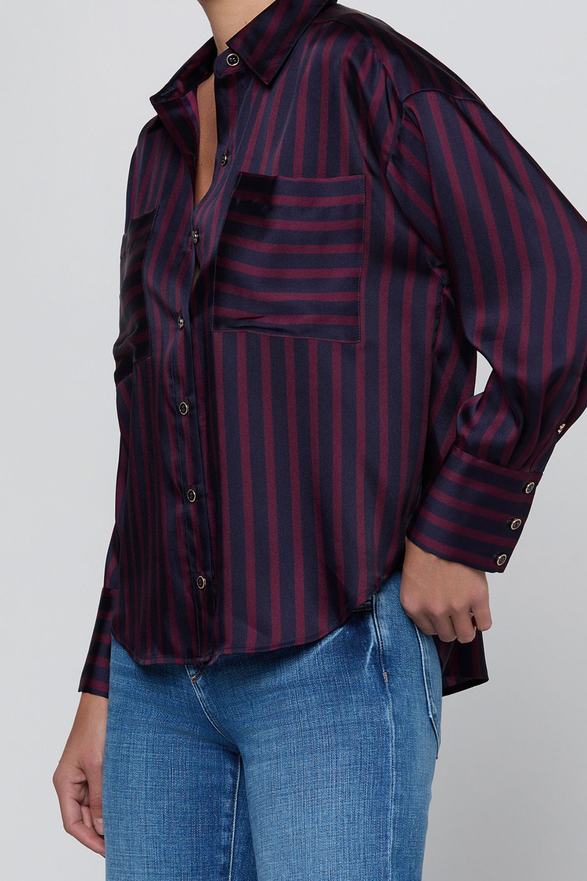 A woman wearing a dark purple and navy blue vertically striped button-up shirt with a chest pocket, paired with blue jeans, shown from the neck down against a light gray background.