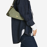 A model wearing a dark denim shirt and jeans, carrying a green fabric shoulder bag with a dark strap and gold-tone hardware against a light gray background.