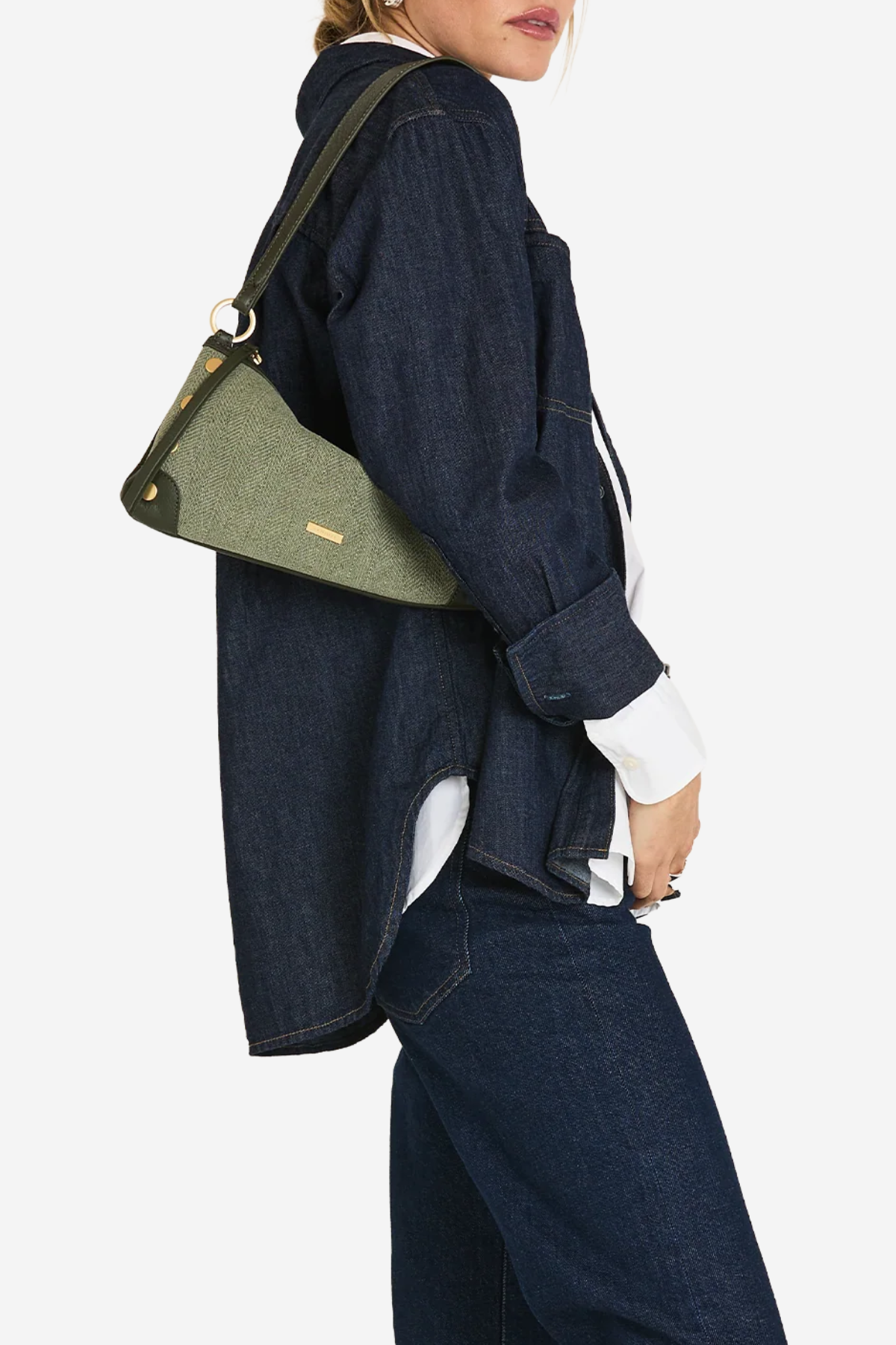 A model wearing a dark denim shirt and jeans, carrying a green fabric shoulder bag with a dark strap and gold-tone hardware against a light gray background.