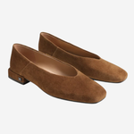 A pair of women's flat shoes in brown suede, featuring a low block heel and a small metallic brand plaque on the outer side, set against a plain light gray background.