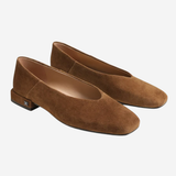A pair of women's flat shoes in brown suede, featuring a low block heel and a small metallic brand plaque on the outer side, set against a plain light gray background.