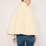 A woman shown from the back wearing a cream-colored cape-style jacket or duster over a white top and blue jeans, photographed against a plain light background.