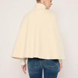 A woman shown from the back wearing a cream-colored cape-style jacket or duster over a white top and blue jeans, photographed against a plain light background.