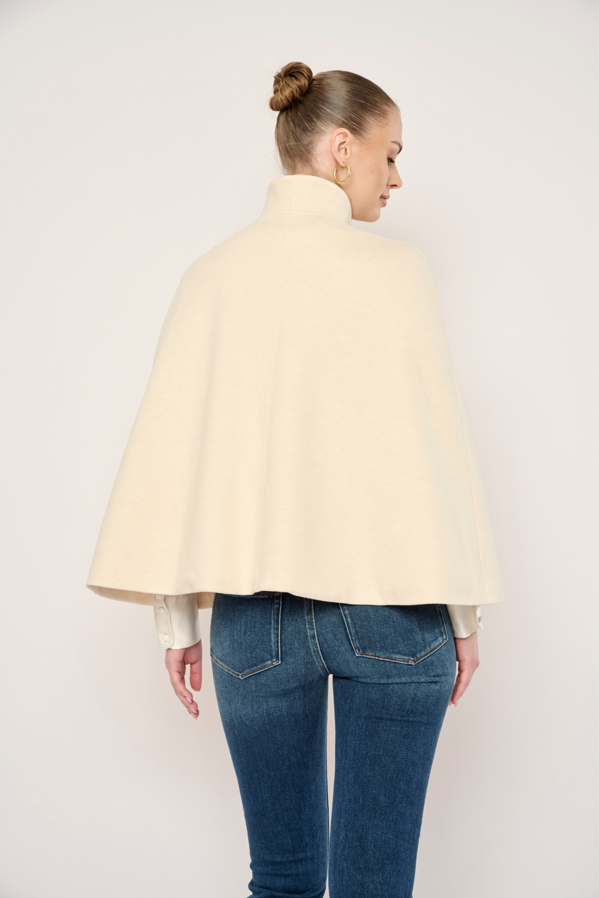 A woman shown from the back wearing a cream-colored cape-style jacket or duster over a white top and blue jeans, photographed against a plain light background.
