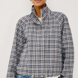 A woman wearing a navy, white, and gray plaid long-sleeve jacket with a high stand-up collar, paired with light blue jeans, against a plain light gray background.