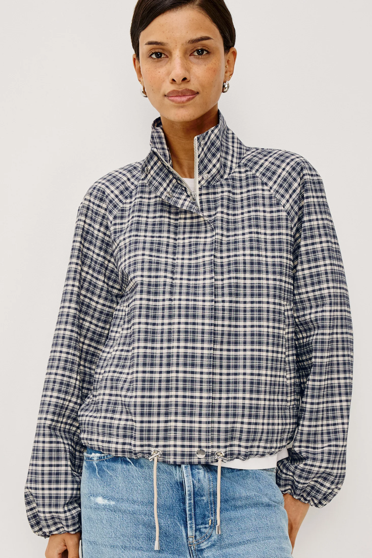 A woman wearing a navy, white, and gray plaid long-sleeve jacket with a high stand-up collar, paired with light blue jeans, against a plain light gray background.